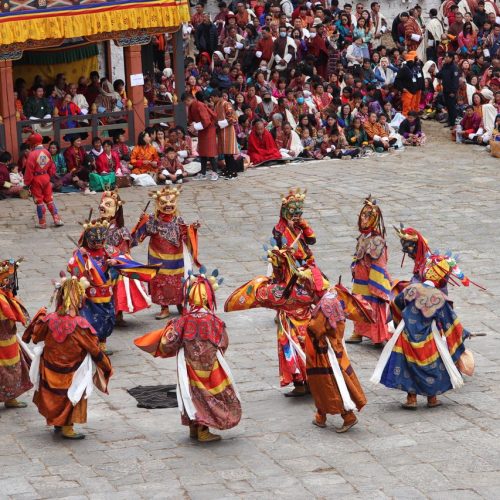 Colorful masked dancers performing at a traditional Bhutanese festival, surrounded by a large crowd.
