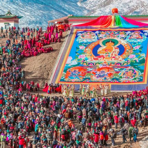 Aerial view of a vibrant Tibetan Buddhist festival with a large thangka and a crowd of participants in colorful robes.